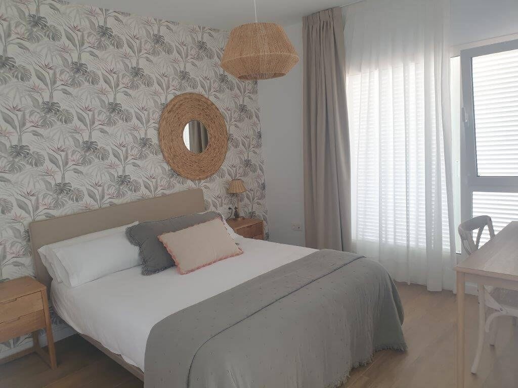Un dormitorio con cama, espejo redondo, mesita de noche, silla y ventana con cortinas.
