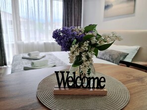 A welcome sign on a woven placemat in a hotel room with a bed, pillows, and a window in the background. - Monfort B (Bansko)