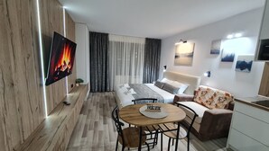 Una habitación de hotel moderna con suelo de madera, una cama con cabecero blanco, un sofá con funda floral, una mesa redonda de madera con posavasos que dice 'Welcome', un televisor montado en la pared y una ventana con cortinas grises.