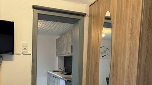 Makuuhuoneessa on sänky, puinen vaatekaappi, seinälle asennettu televisio ja keittiö, joka näkyy avoimen oven läpi.