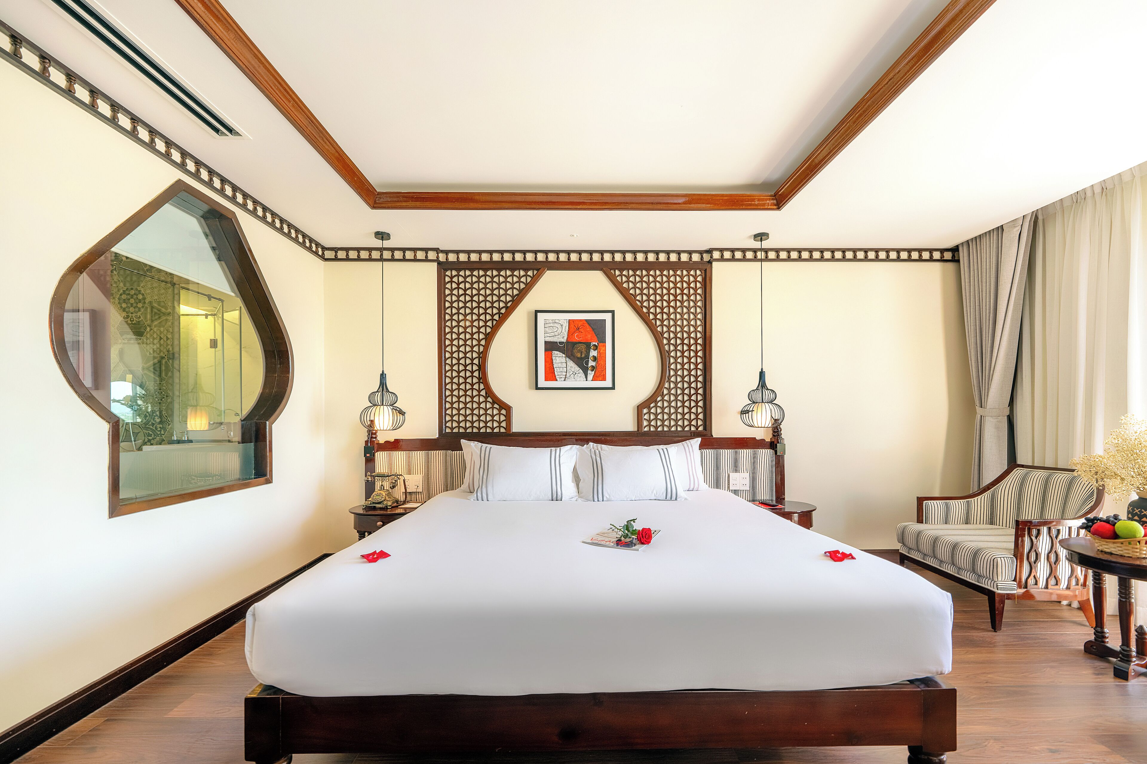 A large bed with white linens, a wooden headboard, and a decorative wall panel. There is a chair with a striped cushion and a small table with fruit.