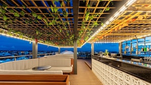 A rooftop bar with a wooden deck, cushioned seating, and a bar area with a view of the cityscape. - Qualia De Hôtel Hoi An - Rooftop Bar City View (Da Nang)
