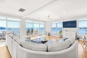 A spacious living area with a large window overlooking the sea, a comfortable sofa, a small coffee table, and a TV screen displaying "OCEANS EDGE VACATIONS".