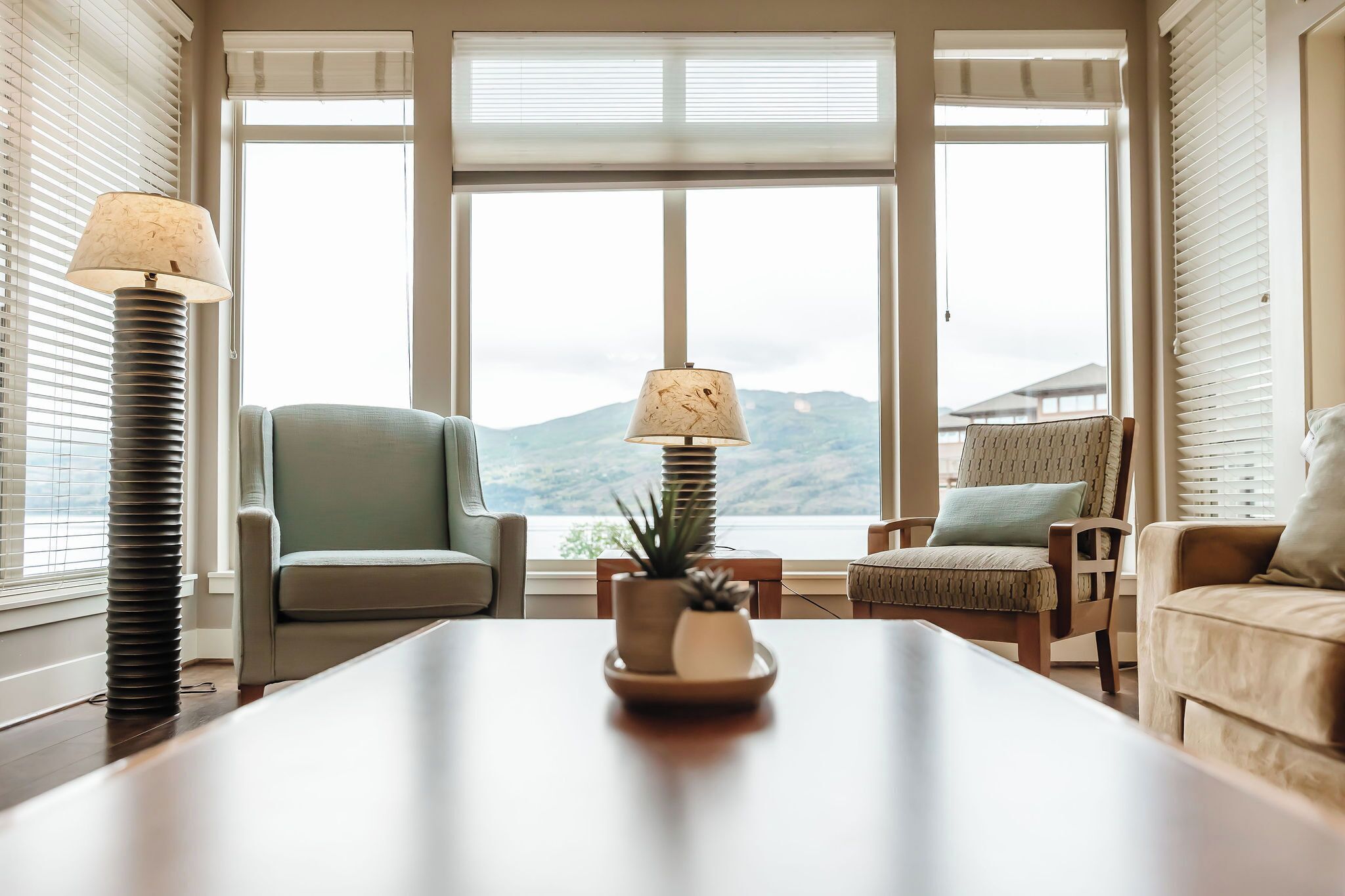 The living room boasts large windows with stunning lake and mountain views. Two armchairs face each other, with a sofa nearby. A coffee table with a potted plant and floor lamps add cozy touches.