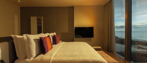 A modern hotel room with a large bed, a desk with a laptop, a TV mounted on the wall, and a large window offering a view of the sea.