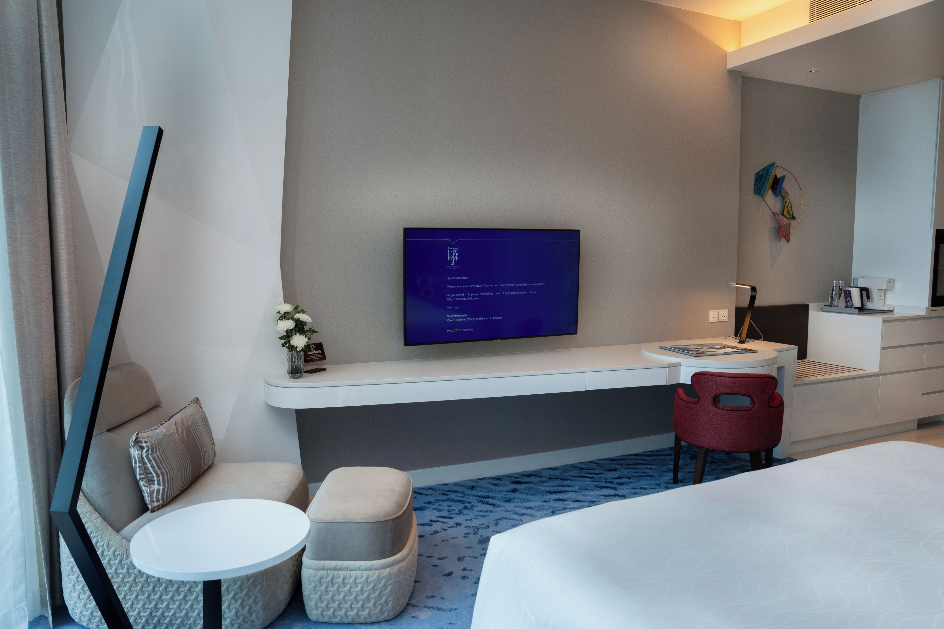 A modern hotel room with a flat-screen TV mounted on the wall, a small round table, a red chair, a bed, and a sofa with a cushion.