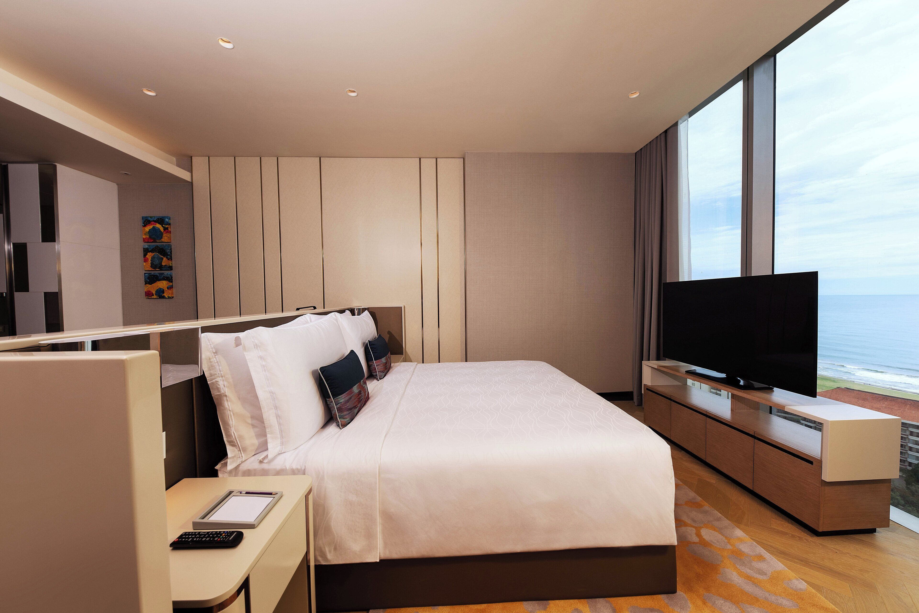 A modern hotel room with a large bed, a flat-screen TV, a desk with a computer, and a view of the ocean.