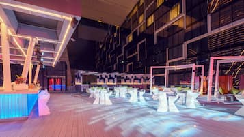 Un espacio interior moderno con suelo de madera, mesas blancas y una zona de bar con iluminación azul.