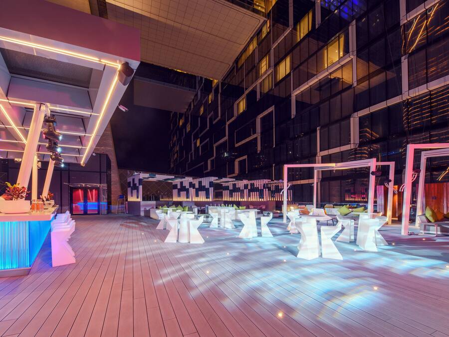 Un espacio interior moderno con suelo de madera, mesas blancas y una zona de bar con iluminación azul.