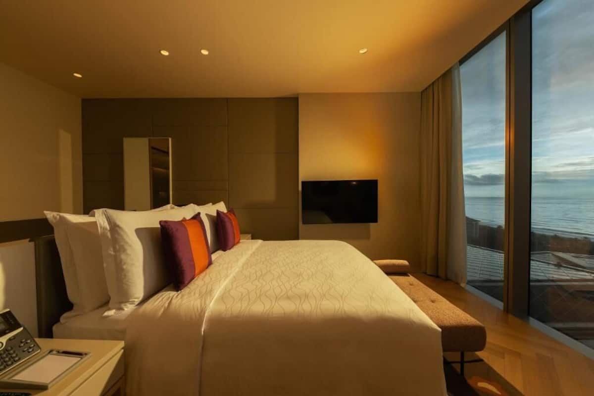 Una moderna camera d'albergo con un letto grande, una scrivania con un portatile, una TV a schermo piatto montata a parete e un'ampia finestra con vista sul mare.