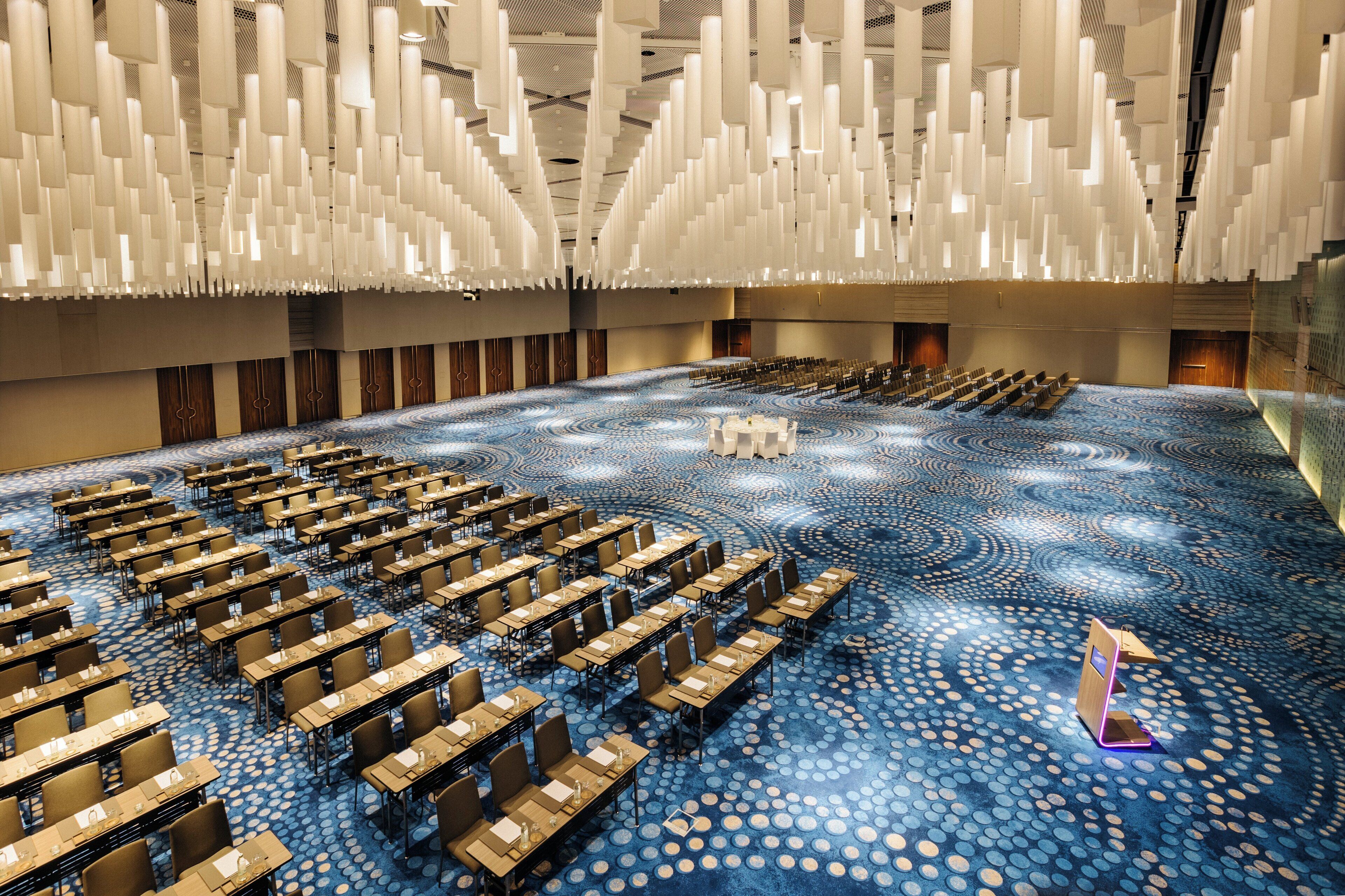 A large conference room with rows of tables and chairs, a patterned carpet, and hanging light fixtures.