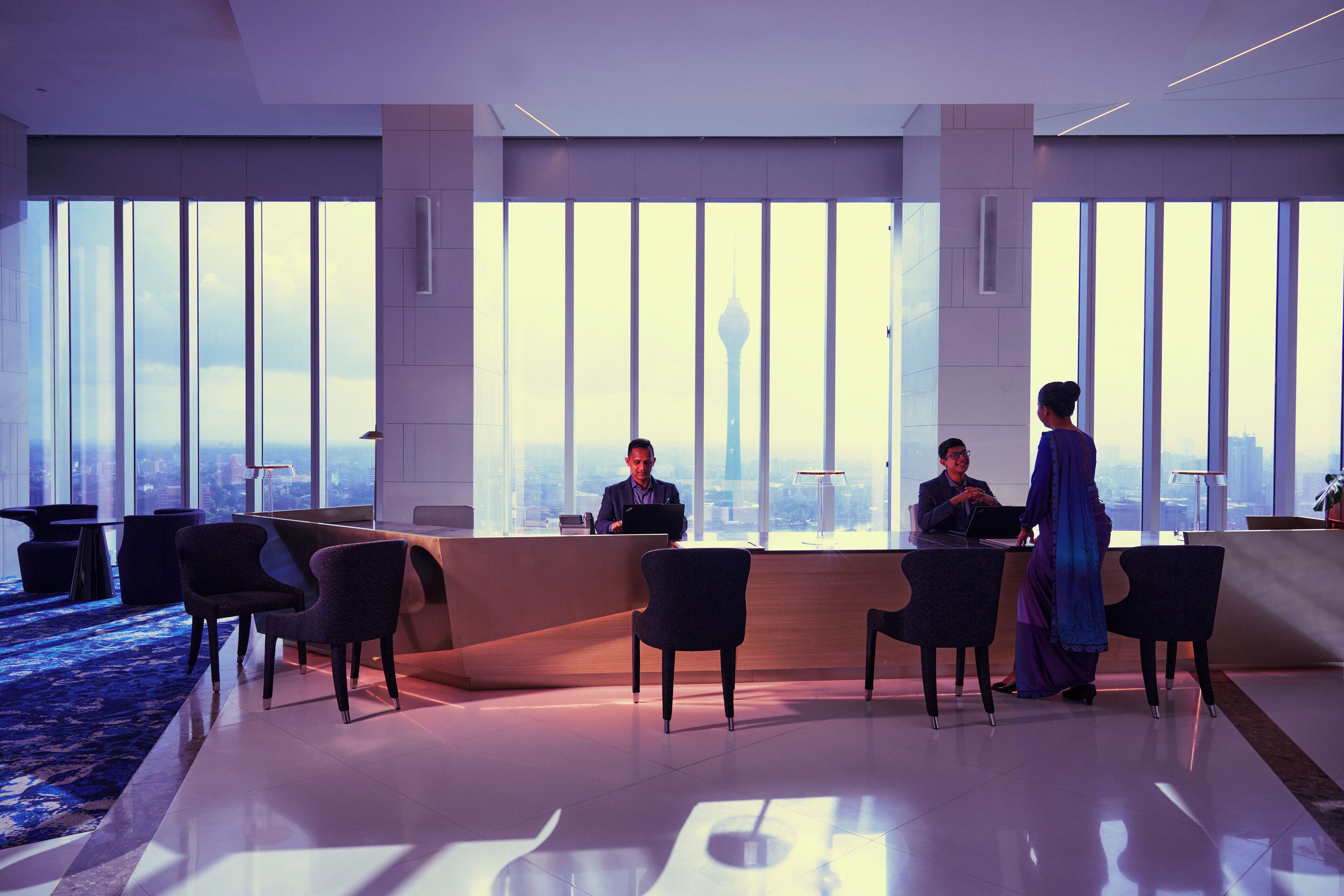 Un área de recepción de hotel moderna con una amplia ventana que ofrece vistas a un horizonte urbano y una torre. Se observan mostradores de recepción, varias sillas y una zona alfombrada en azul.