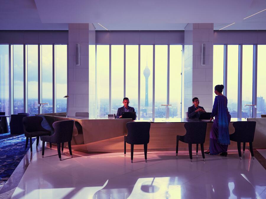 A modern hotel reception area with a large window view of a city skyline and a tower. There are reception desks, several chairs, and a blue carpeted area.