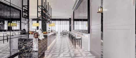 Un moderno vestíbulo de hotel con piso de diseño geométrico, un área central de asientos con mesas y sillas, y un bar con estante alto que exhibe botellas.