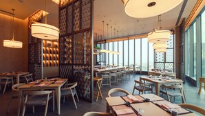 Un restaurante moderno con varias mesas y sillas, ventanas amplias y luminarias decorativas.
