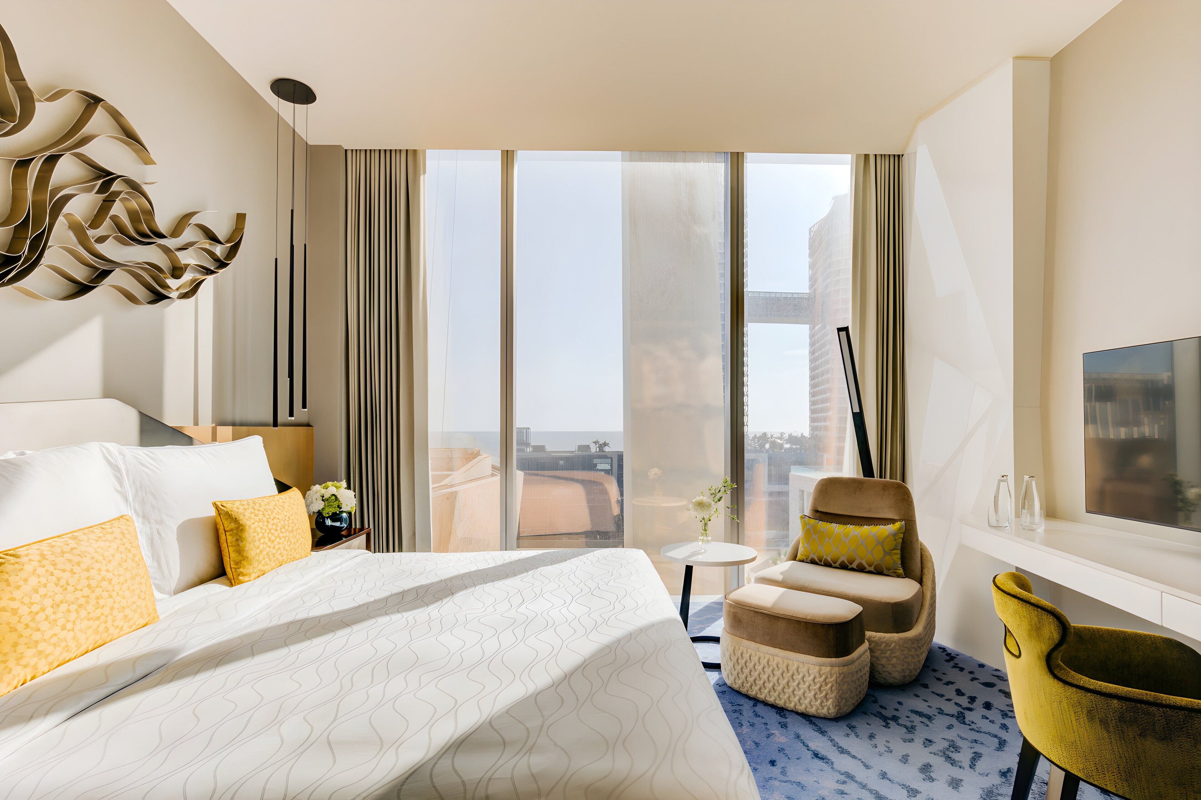 Une chambre d’hôtel dotée d’un grand lit, d’une télévision, d’un coin salon avec des fauteuils et offrant une vue sur la ville grâce à de grandes fenêtres.