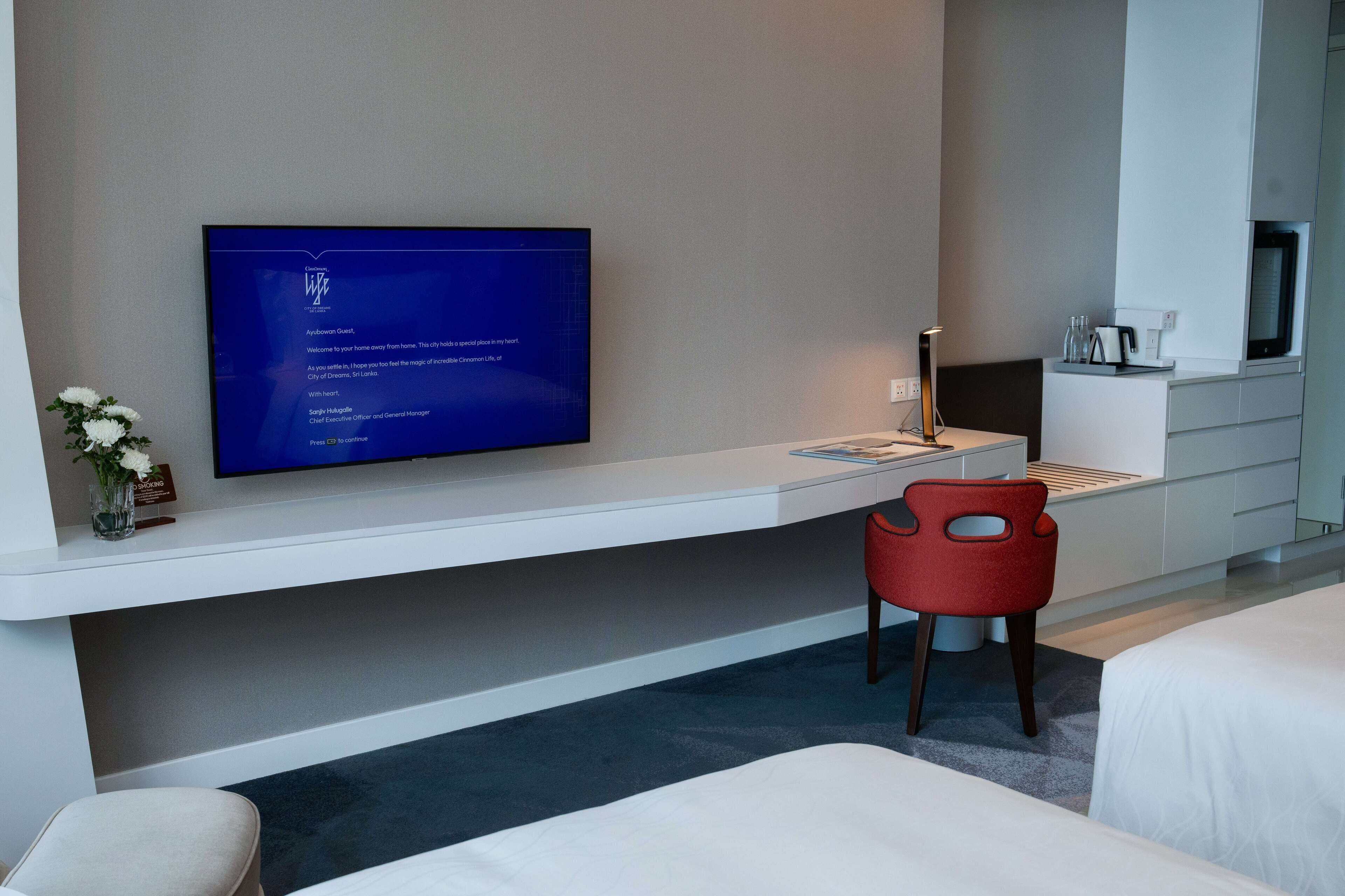 Une chambre d’hôtel équipée d’un téléviseur à écran plat fixé sur une console blanche, d’un fauteuil rouge, d’un lit et d’une petite table avec une lampe.
