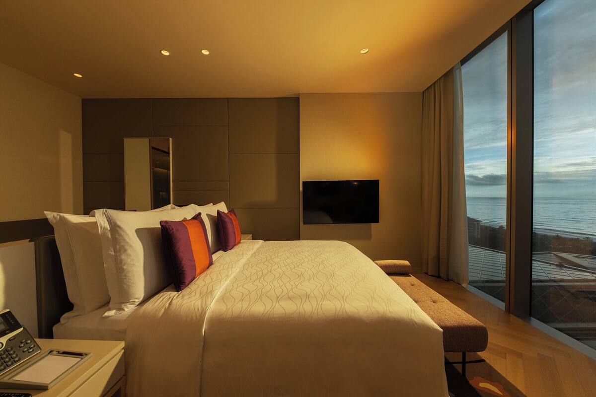 Una moderna camera d'albergo con un letto grande, una scrivania con un portatile, una TV a schermo piatto fissata al muro e un'ampia finestra con vista sull'oceano.
