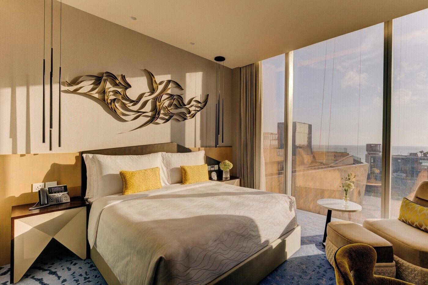A modern hotel room with a large bed, a bedside table with a phone, a small table with a vase, and a view of the city through large windows.