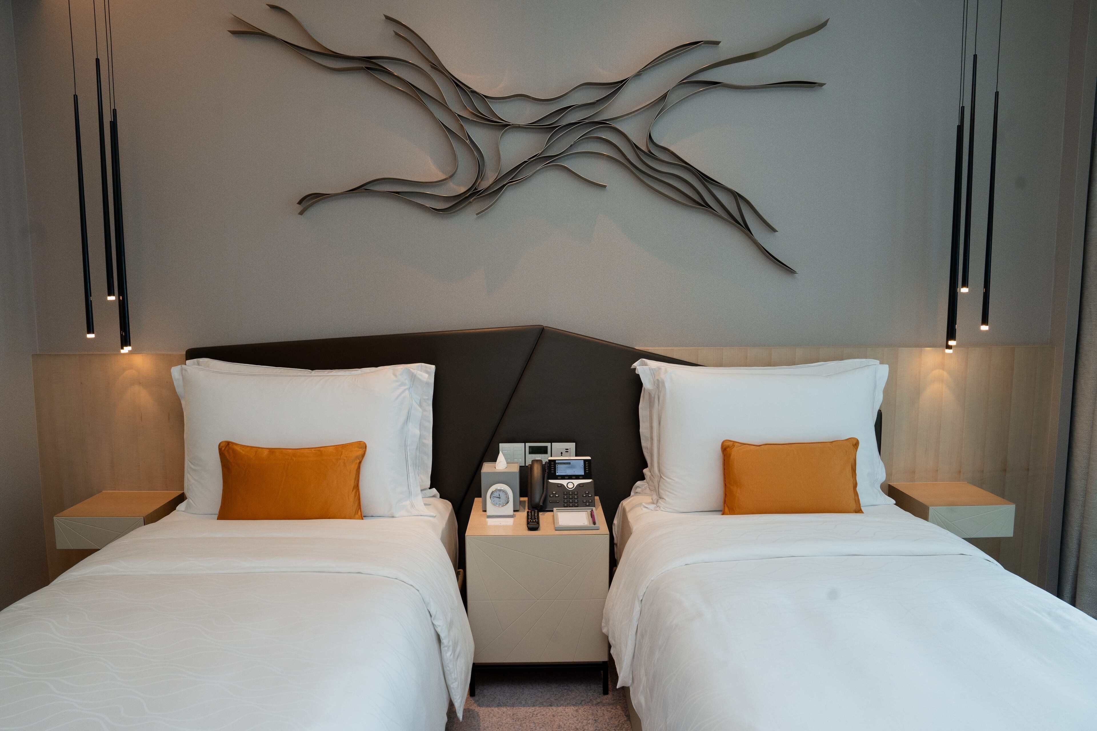 Una habitación de hotel moderna con dos camas, una obra de arte colgada en la pared, lámparas de noche y una mesita de noche con teléfono y reloj.