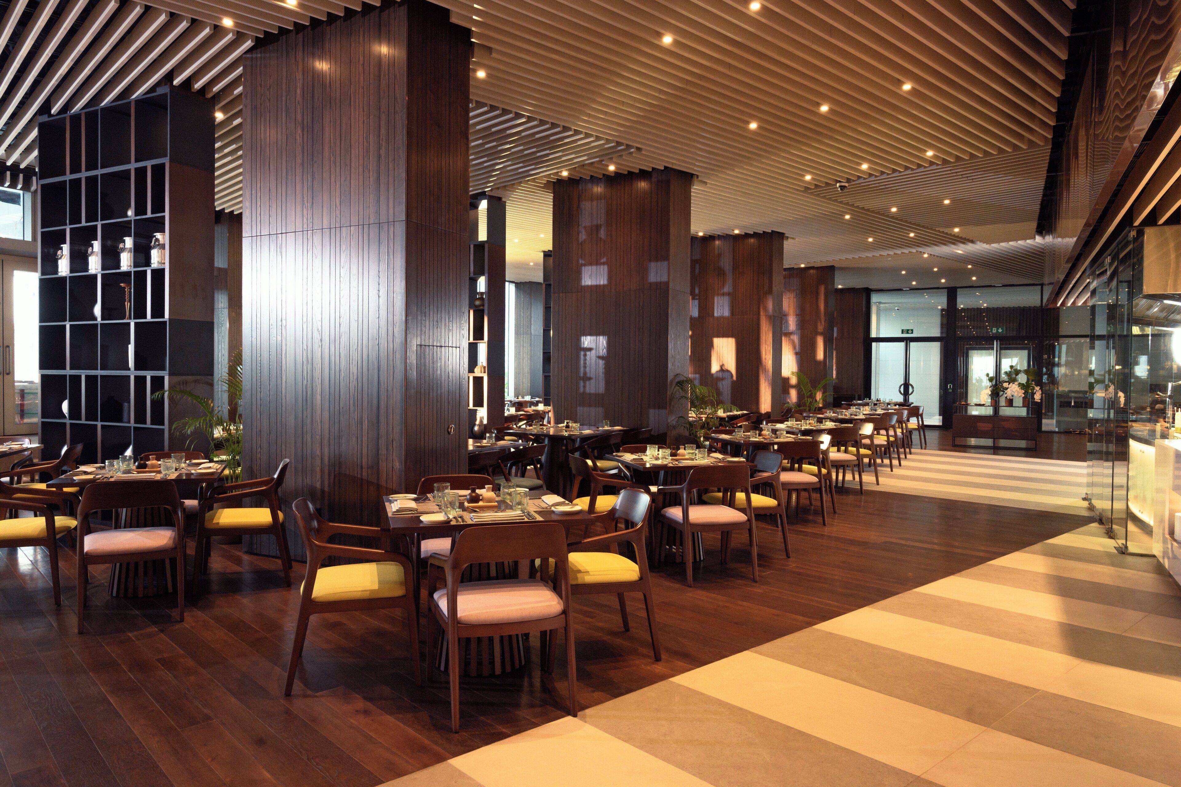 A spacious dining area with multiple tables and chairs, wooden flooring, and a modern ceiling design.