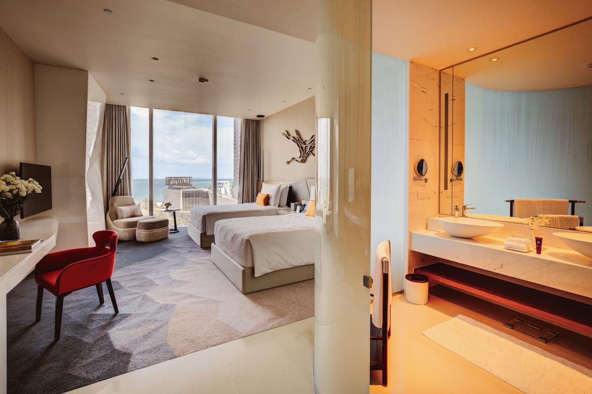 Una camera d'albergo moderna con un letto grande, una scrivania con una sedia, un bagno con lavandino e specchio, e vista sull'oceano.