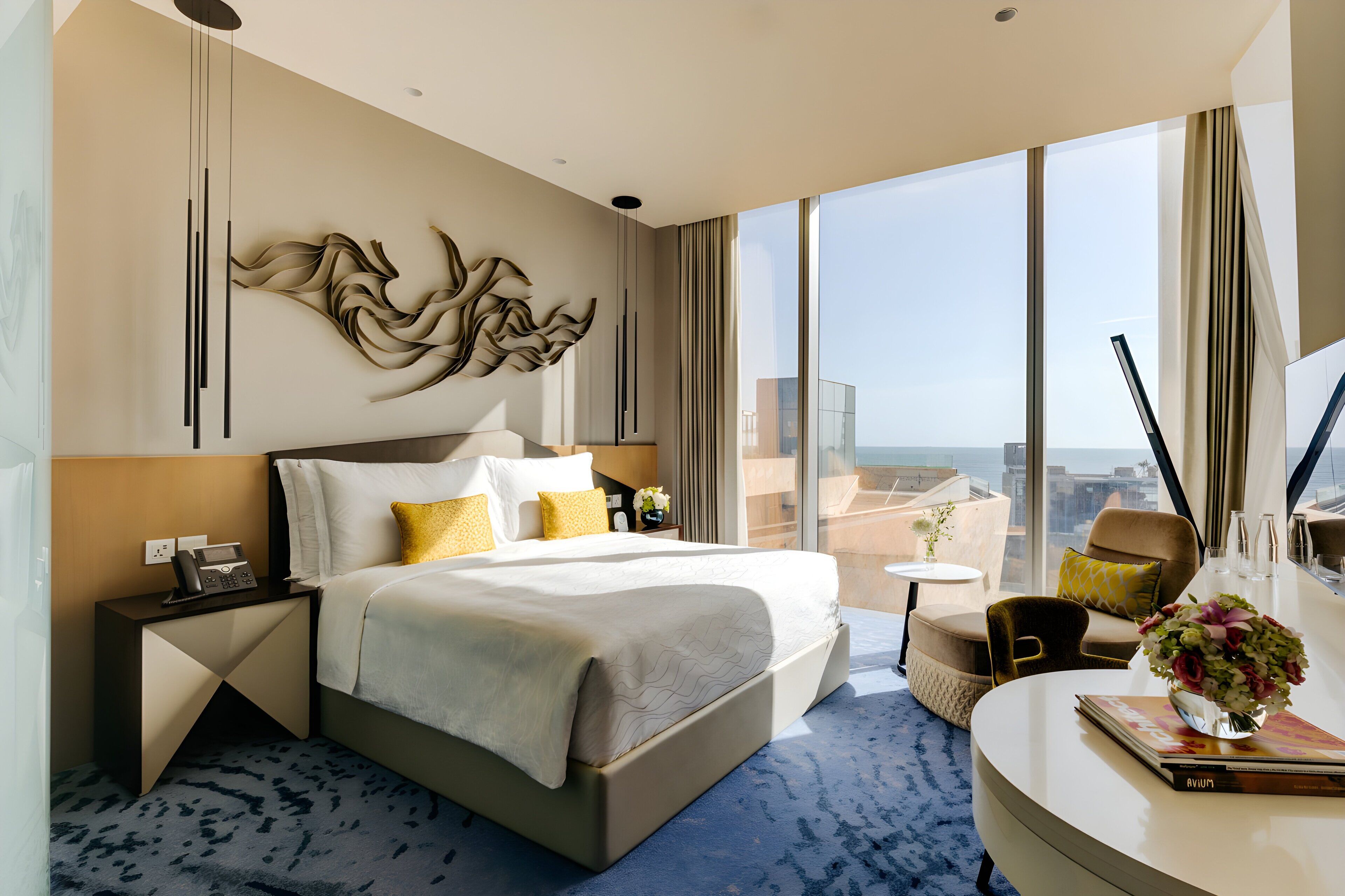 Quarto de hotel moderno com uma cama grande, mesa de cabeceira com telefone, pequena mesa com um vaso e vista da cidade através de grandes janelas.