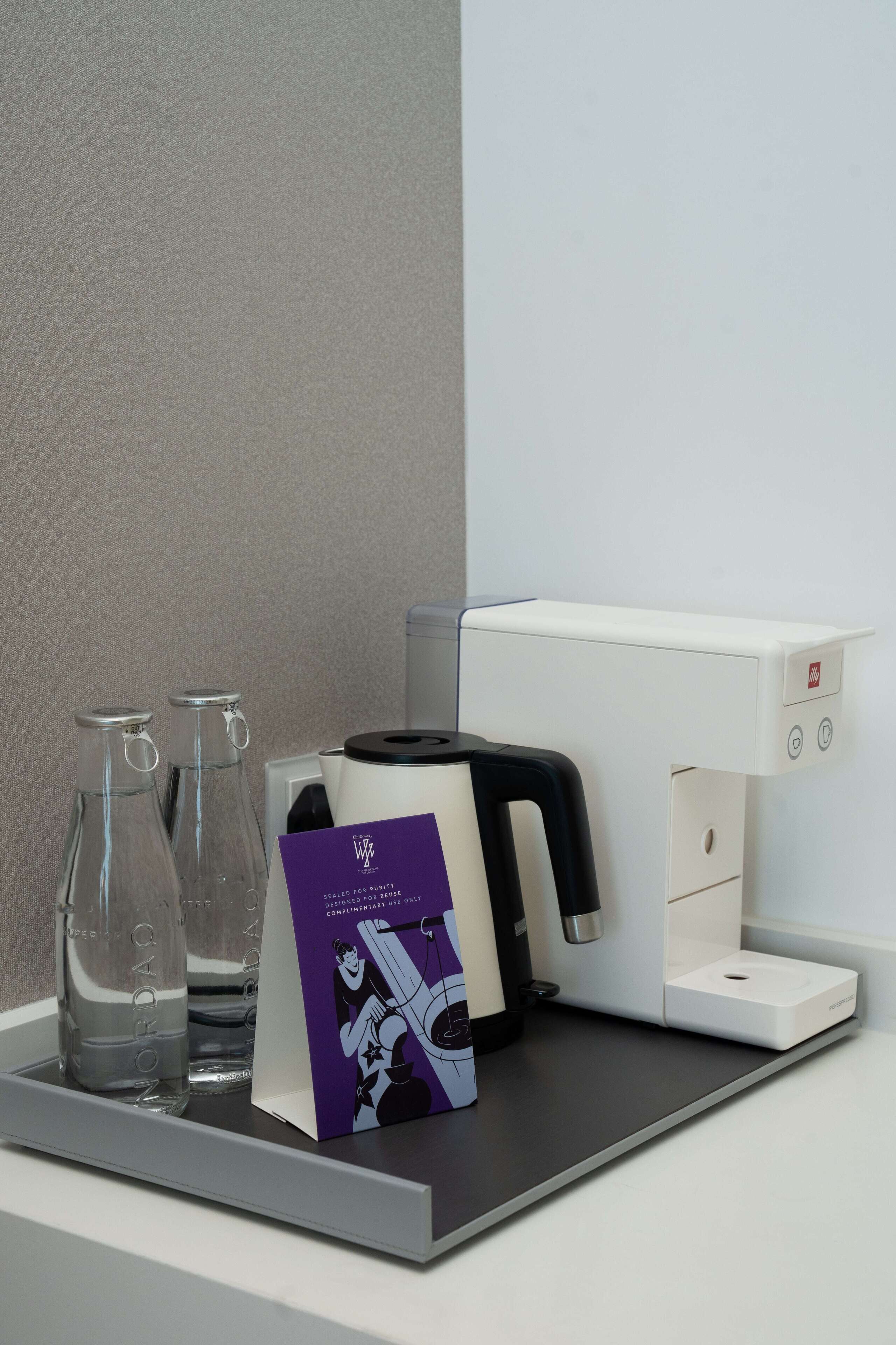 A hotel room's minibar with a coffee machine, two glass bottles, and a purple brochure.