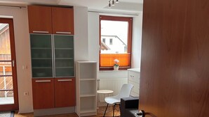 Ein Zimmer mit Holzboden, einem Schrank mit Glastüren, einem Heizkörper, einem kleinen Tisch mit Stuhl und einem Fenster mit Jalousien.