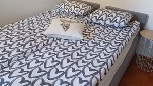 Ein ordentlich bezogenes Bett mit einem zudeckenen Bettbezug mit Herzmuster und passenden Kissen, ein kleiner runder Beistelltisch und ein an der Wand hängendes Bild.
