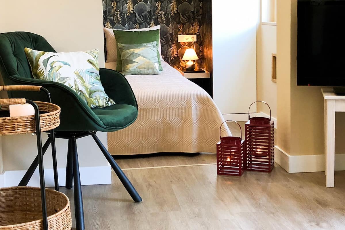 Een moderne slaapkamer met een groene stoel, een bed met gekleurde kussens, een nachtkastje met een lamp, een televisie en rieten manden.