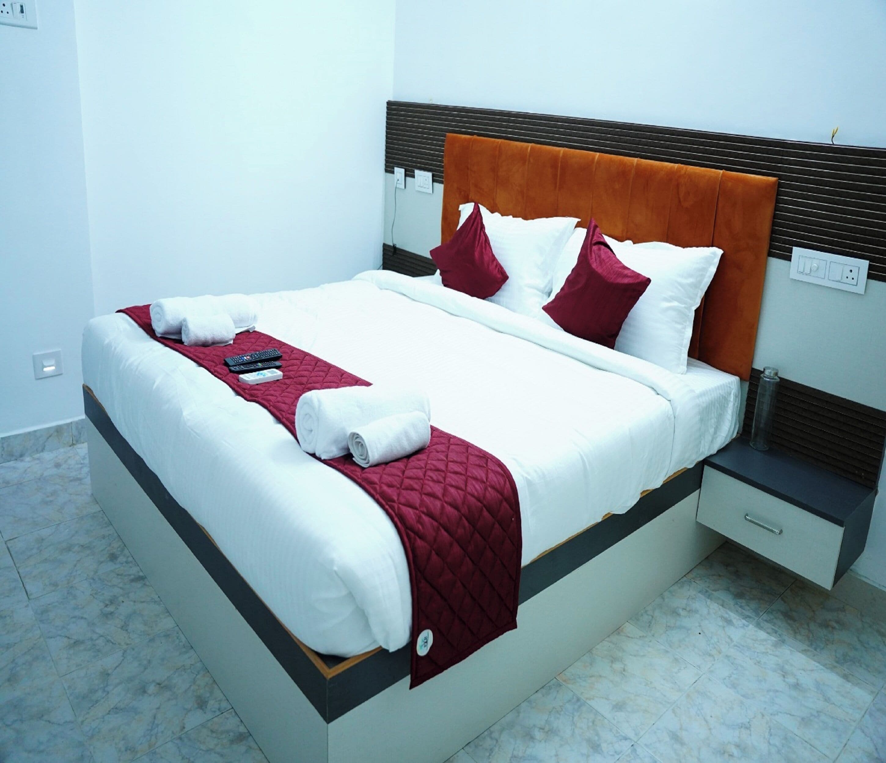 Een net opgemaakt bed met wit en rood beddengoed, een gerimpelde rode deken en twee afstandsbedieningen op het bed.