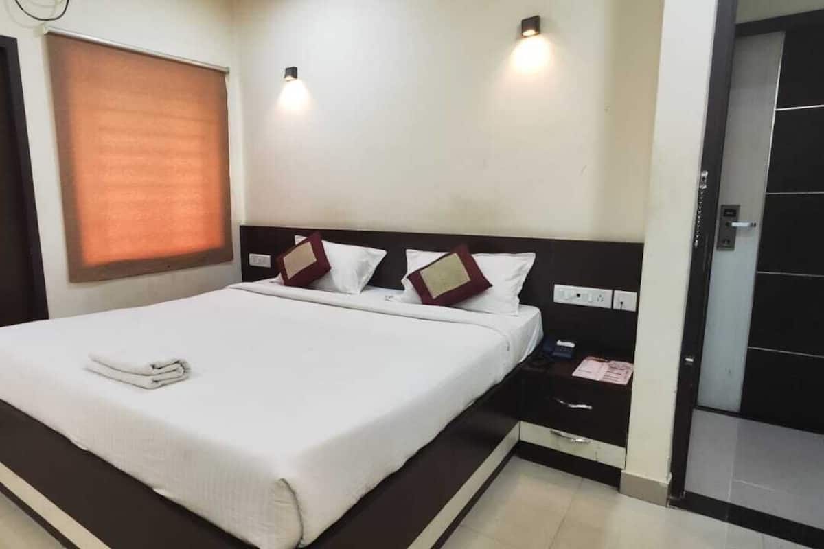 A hotel room with a bed, a bedside table, a nightstand, a wall-mounted light, a window with blinds, and a door with a number plate.