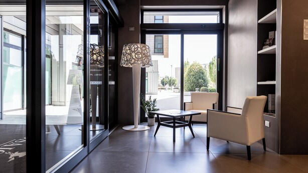 A modern interior with a glass door leading outside, a white chair, a small table, and a decorative lamp. - Mosaico Suite (Cesena)
