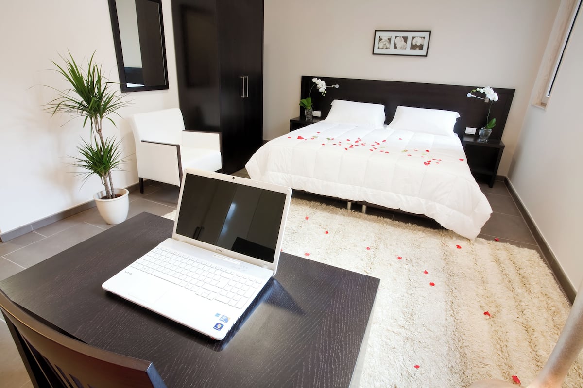 Ein modernes Hotelzimmer mit einem Laptop auf einem Schreibtisch, einem Bett mit Rosenblättern, einer Topfpflanze und einem weißen Stuhl.