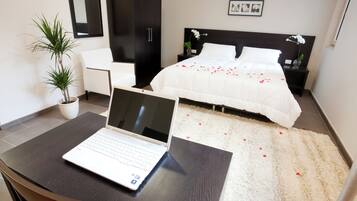 Una habitación de hotel moderna con un ordenador portátil sobre un escritorio, una cama decorada con pétalos de rosa, una planta en maceta y una silla blanca.