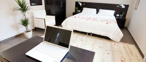 A modern hotel room with a laptop on a desk, a bed with rose petals, a potted plant, and a white chair.
