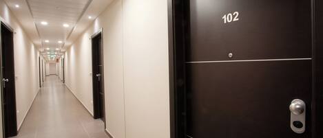 Ein Hotelkorridor mit langer, schmaler Gestaltung, einer Tür mit der Beschriftung „102“ und einem Fluchtwegweiser an der Decke.