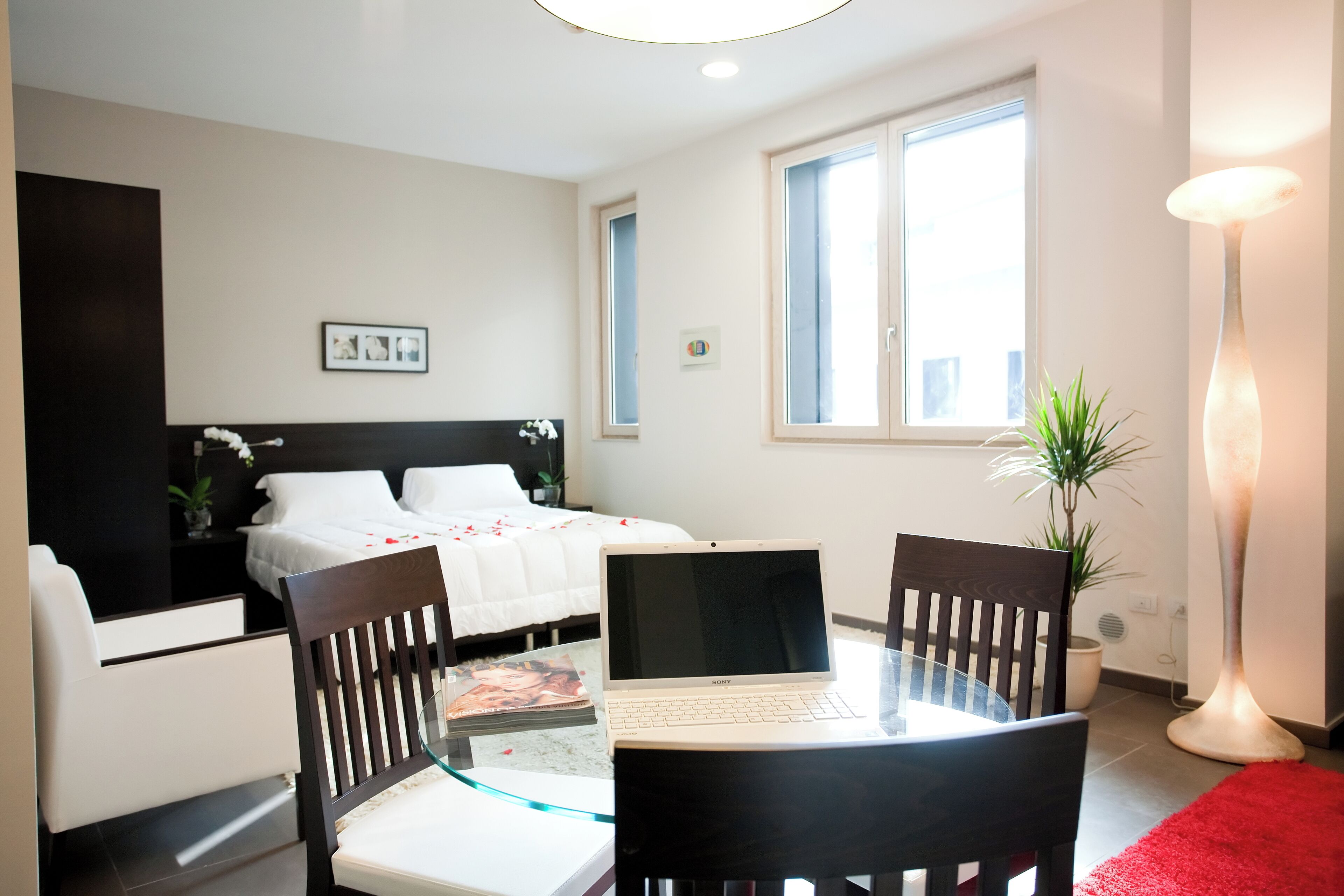 A modern hotel room with a bed, a desk with a laptop, a chair, a red rug, and a floor lamp.