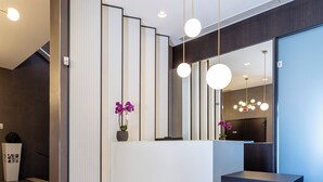 A modern reception desk with a large mirror, vertical panels, and hanging pendant lights. - Mosaico Suite (Cesena)
