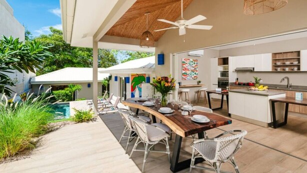 Un espace repas extérieur moderne, avec une table en bois et des chaises en osier, un ventilateur de plafond, et une cuisine dotée de meubles blancs et d’un plan de travail en bois.