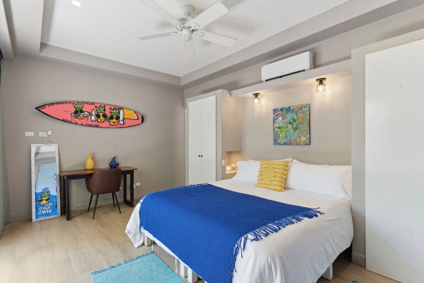 A bedroom with a bed, a desk, a chair, a ceiling fan, and artwork on the walls.