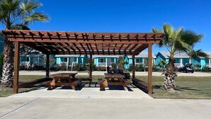 Eine hölzerne Pergola mit Picknicktischen und Bänken, umgeben von Palmen und einer Reihe blauer Häuser.