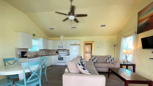A modern living room with a beige sofa, a dining area with turquoise chairs, a kitchen with white cabinets, and a ceiling fan.