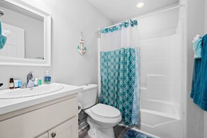 Une salle de bain avec un lavabo, des toilettes et une baignoire, tous de couleur blanche. Il y a un rideau de douche orné d’un motif géométrique.