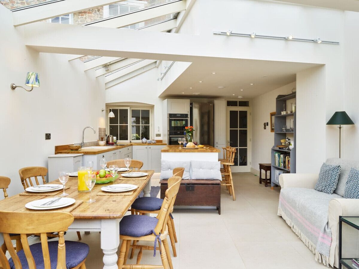 Een ruime woonkamer met een eettafel voor vier personen, een keuken op de achtergrond en een gezellige zithoek met een bank en een vloerlamp.