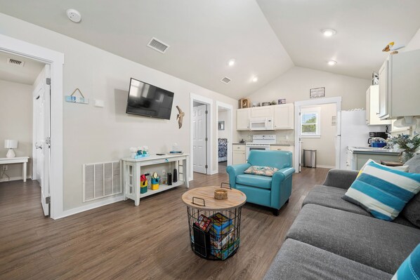 A modern living room with a grey sofa, a blue armchair, and a round wooden table. There is a TV mounted on the wall, a white cabinet, and a small side table with decorative items.