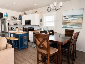 A modern kitchen with white cabinets, a microwave, and a coffee machine. There is a dining area with a wooden table and chairs.