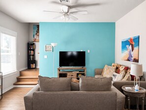 A living room with a teal wall, a flat-screen TV, a ceiling fan, a bookshelf, and a staircase.