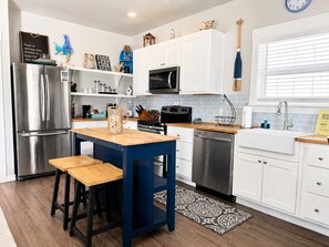 A modern kitchen with a stainless steel refrigerator, a microwave, a stove, and a sink. There is a wooden table with two stools.